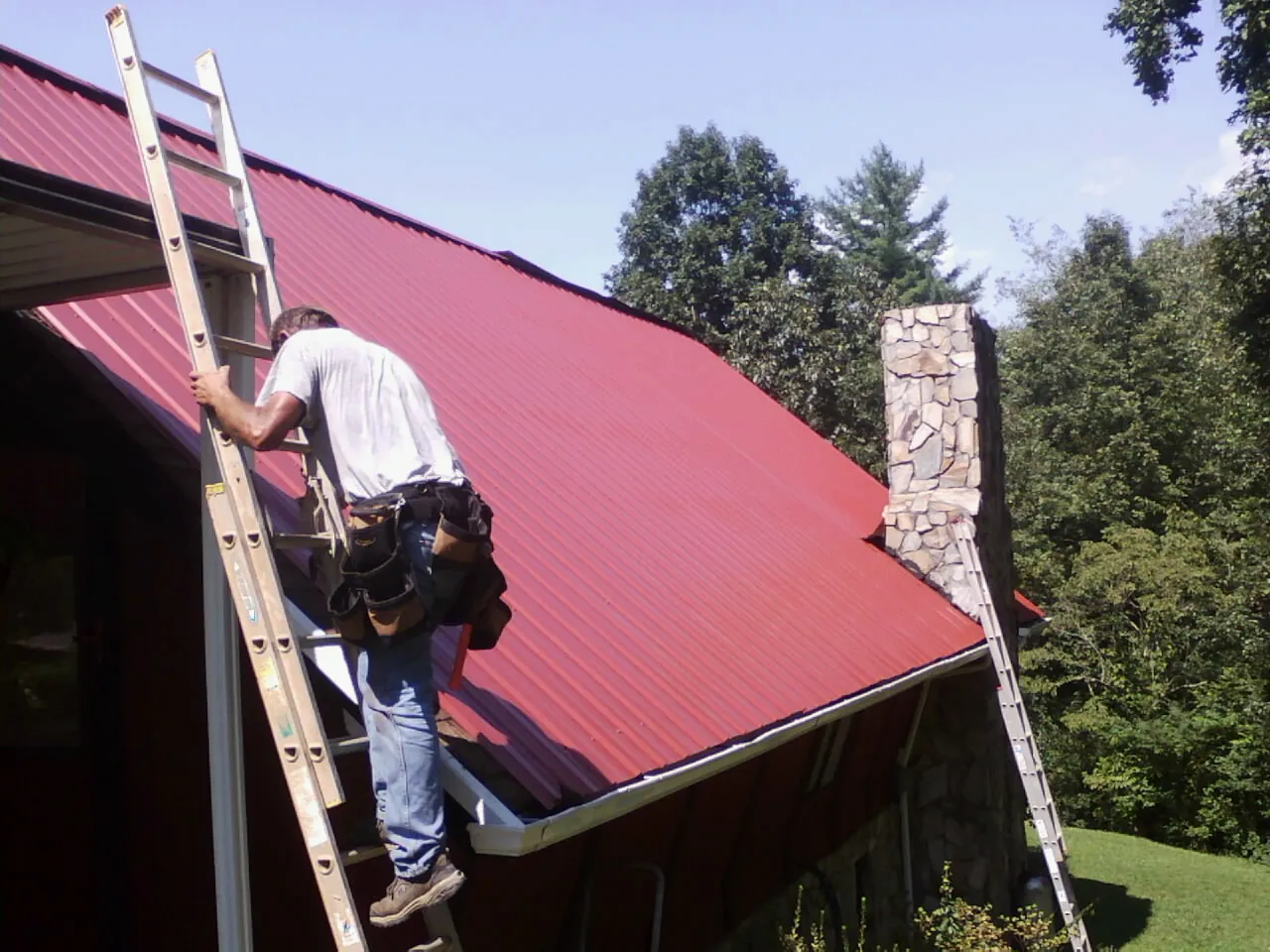 Roofer on a ladder beside a red metal roof and stone chimney — Soffit & Fascia Repair by Roofing Brothers in Center Hill
